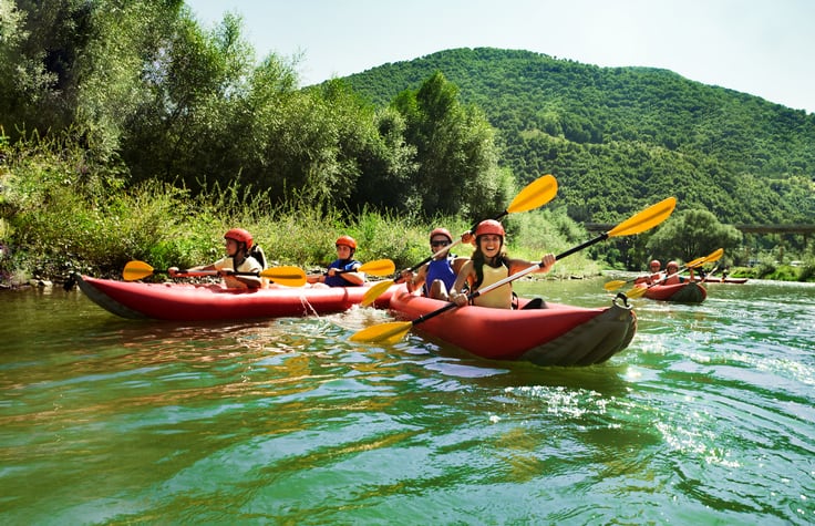 Several teams of two go rafting in inflatable canoes on calm waters in a magnificent setting.