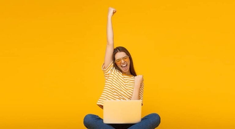 Una mujer joven levanta el brazo delante de su ordenador, parece estar jugando a un juego y haber ganado. La pared detrás de ella es amarilla.