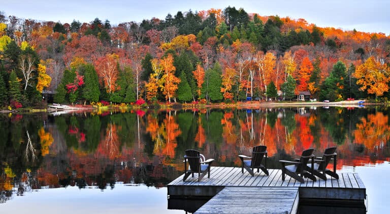 Un lago en un día de otoño con un pontón y sillas. Los árboles son verdes, naranjas y amarillos.