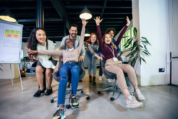 De jeunes hommes d'affaires joyeux en tenue décontractée élégante s'amusent tout en courant sur des chaises de bureau et en souriant.