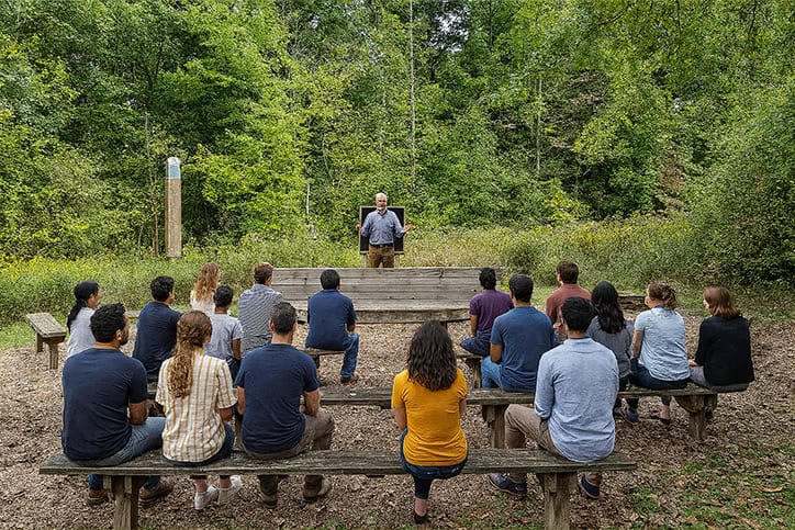 Séminaire en plein air dans une clairière forestière, avec plusieurs personnes assises sur des bancs en bois disposés en arc de cercle, tournées vers un orateur debout sur un petit podium en bois, entouré de verdure et d’arbres feuillus.